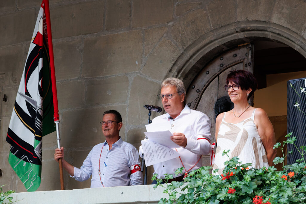 Trois personnes sur un balcon lors d'une cérémonie officielle avec drapeau